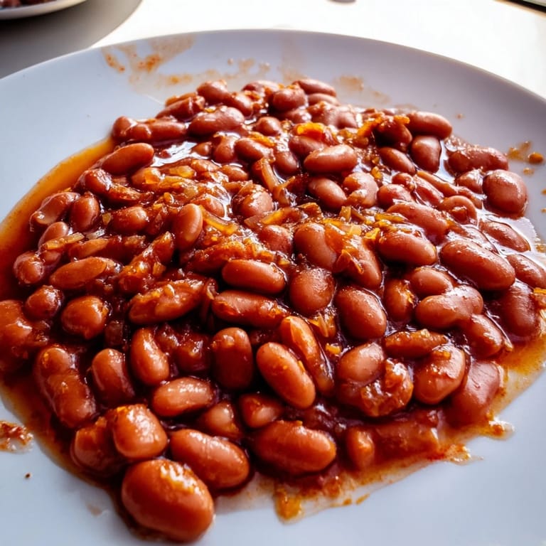 Close-up of glazed Baked Beans topped with fresh parsley, served alongside crusty bread and grilled sausages.